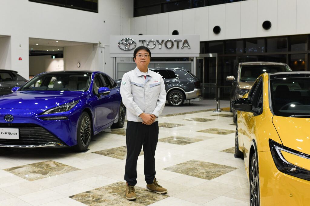 A man stands with his hands clasped in front of him surrounded by gleaming cars.