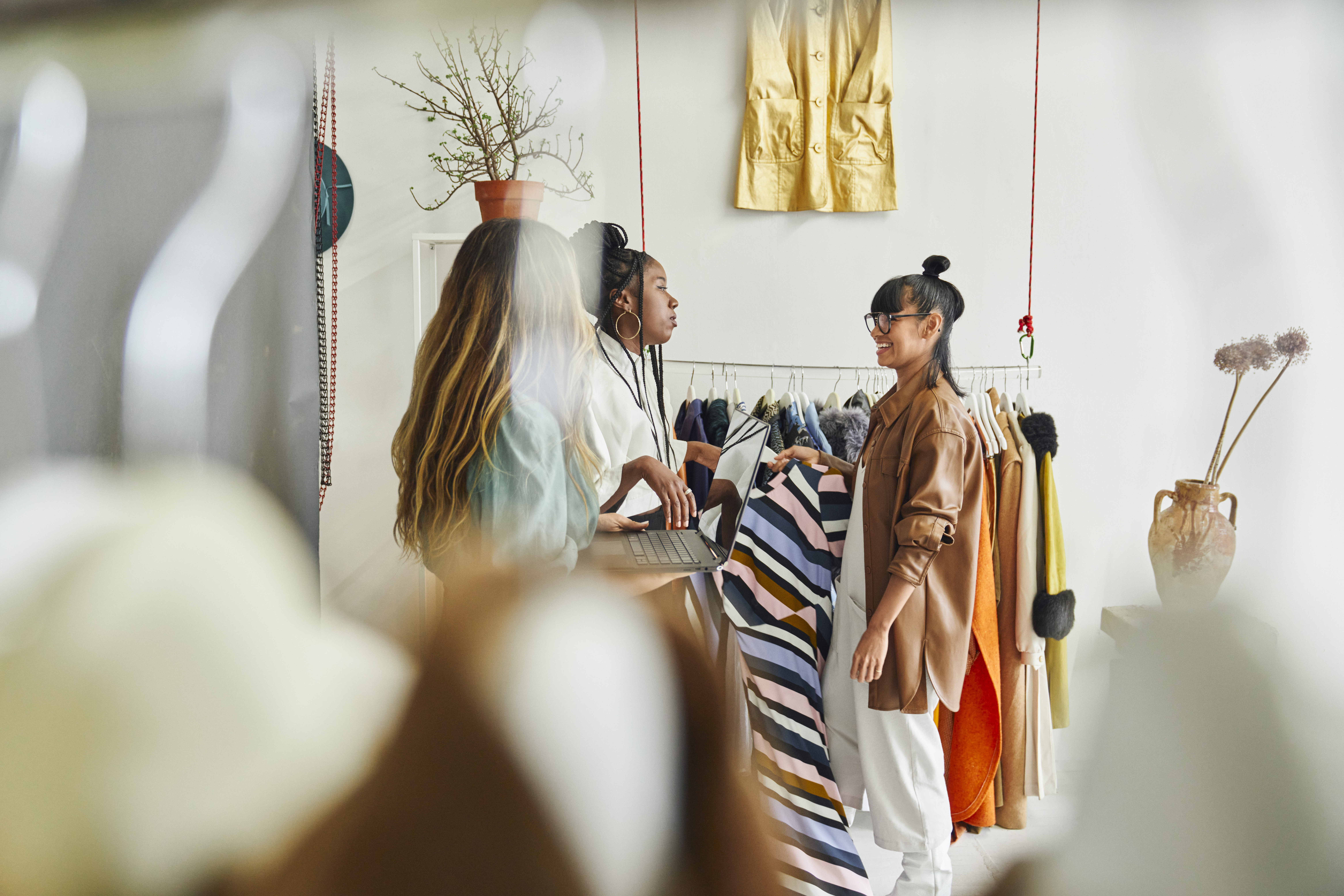 Three people are standing in a clothing store, engaged in conversation. They are surrounded by various garments on hangers and racks, including a striped dress one person is holding. The store has a minimalist decor with plants and hanging clothes.