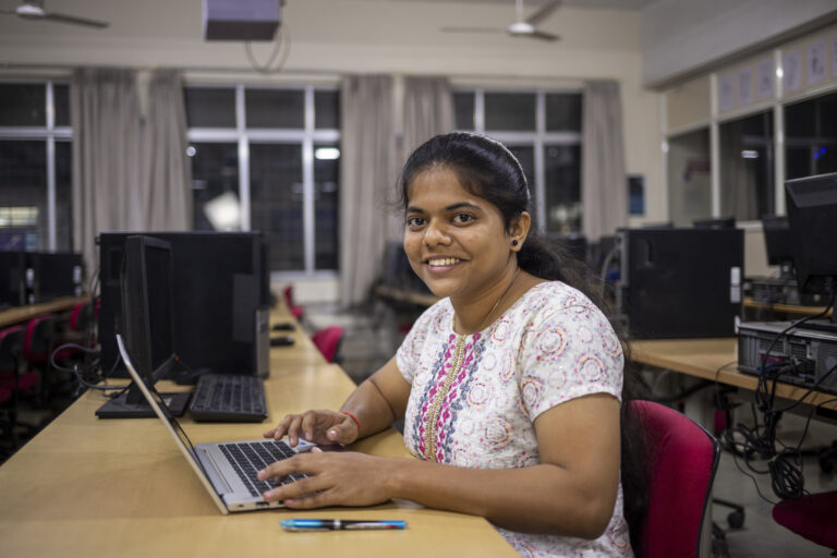 A young woman with her hair tied back sits at a desk with a laptop.