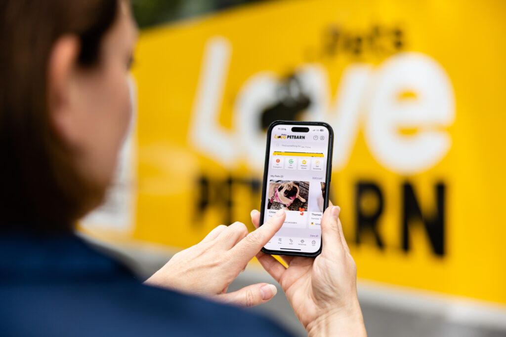 A person is holding a smartphone and interacting with an app on the screen. The background features a large yellow sign with the words 'Love' and 'PETBARN' prominently displayed.