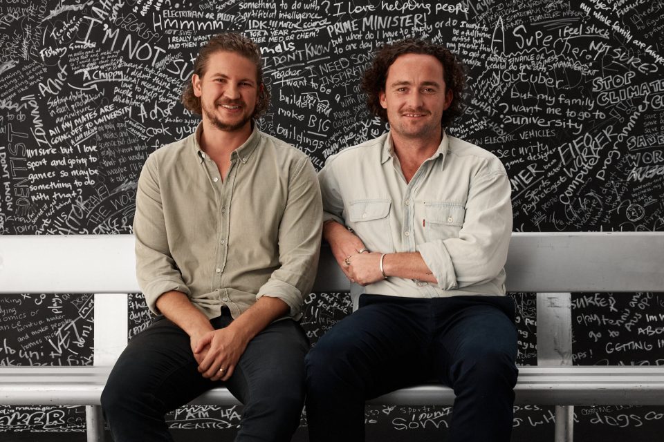 Two adult males sitting on a bench infront of a chalkboard wall filled with texts