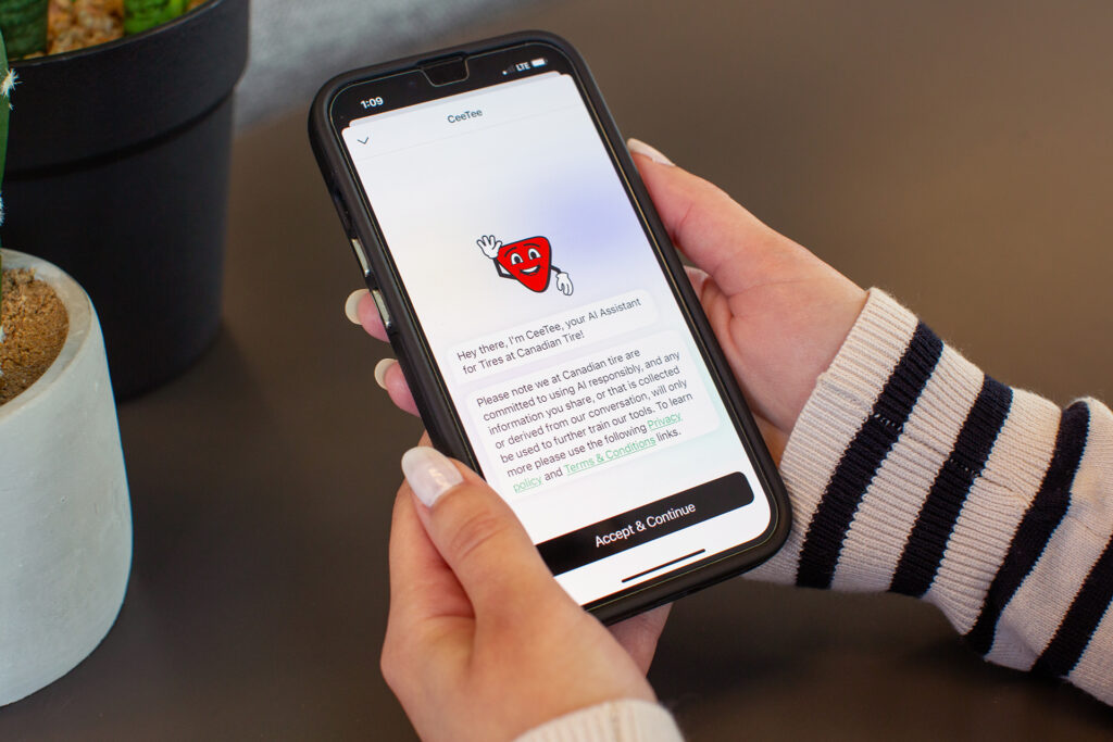 A hand holds a smart phone showing an app with a Canadian Tire logo with text underneath