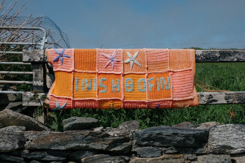 A hand-knitted sign with flowers and patterns in pastel tones spells out “Inishbofin.”
