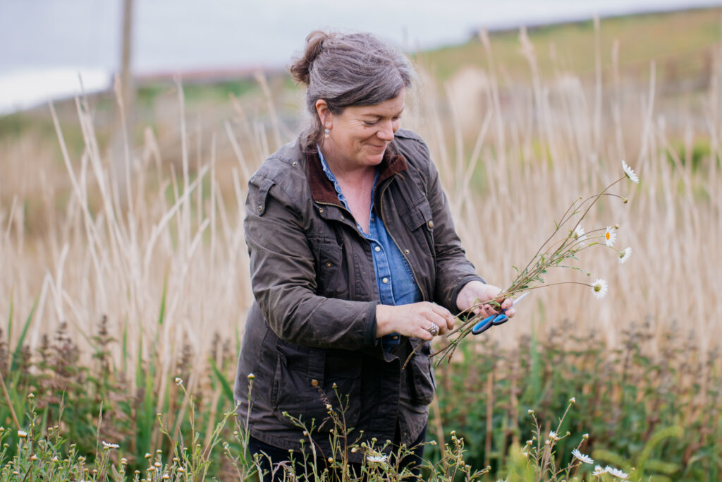 A smiling woman in a jacket gathers flowers in a field of tall grasses and wildflowers. 