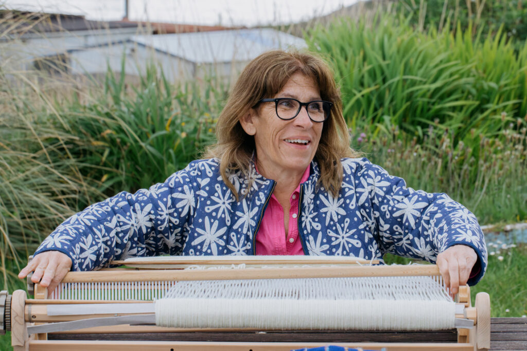 A smiling woman in a blue-and-white floral jacket works at a traditional loom that is flat on a table. 