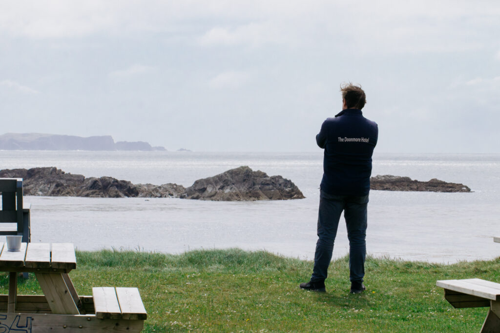 A man in a blue windbreaker with the words “Doonmore Hotel” is portrayed from the back as he gazes out to sea.