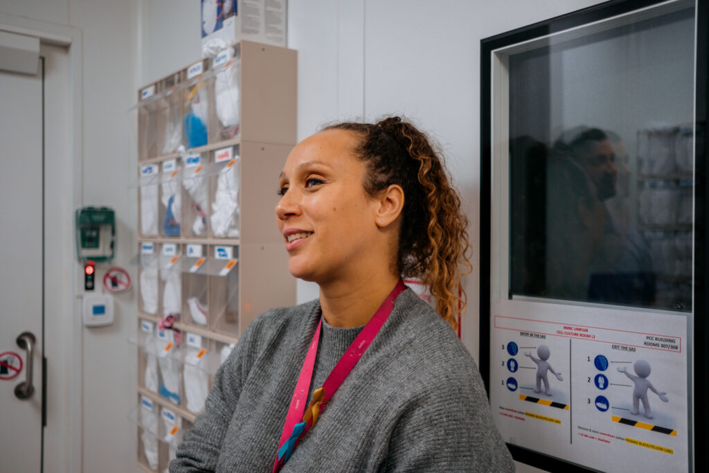 A woman in a gray sweater, with a pink lanyard, in a room with lab supplies.