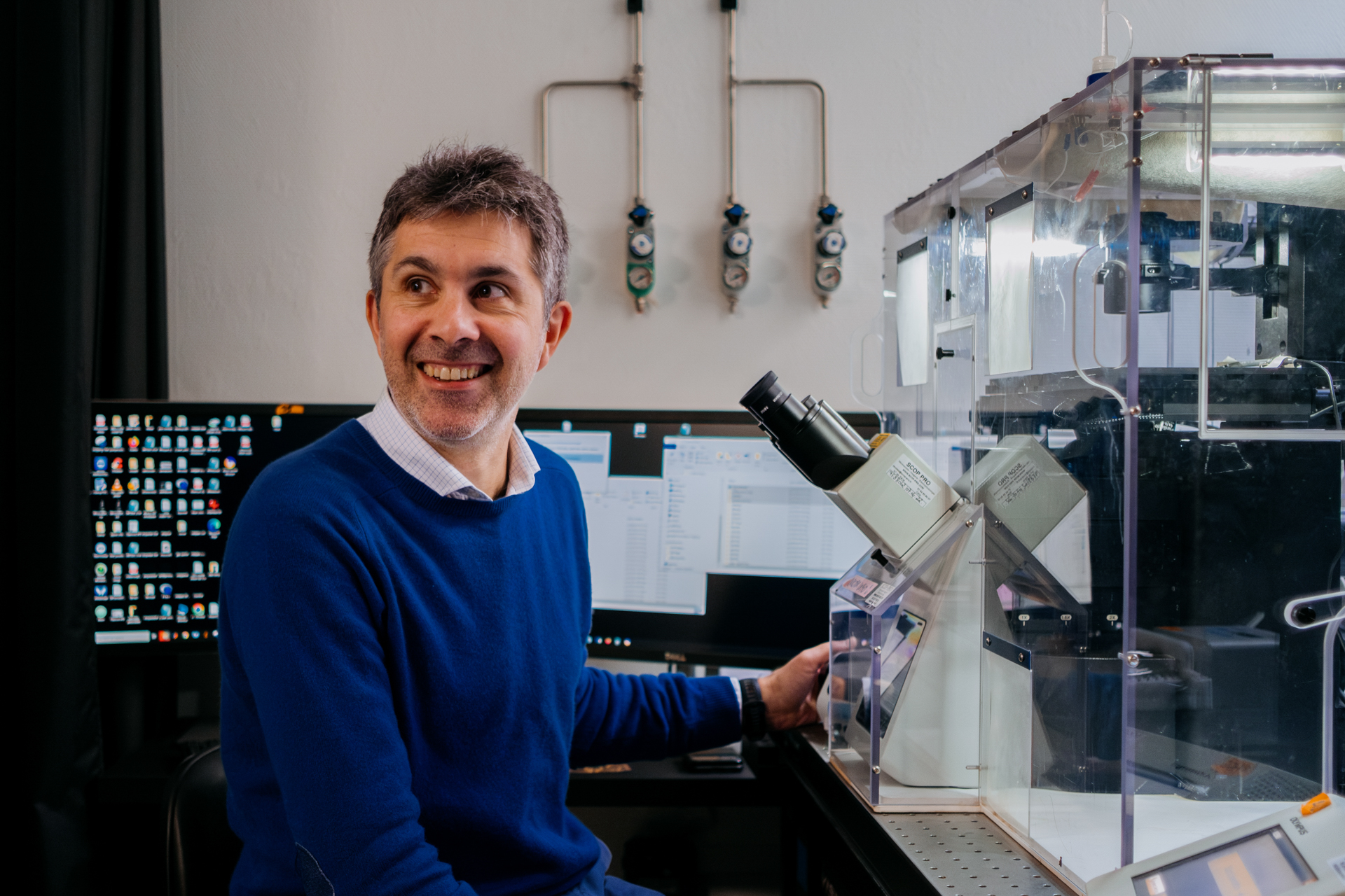 A man in a blue sweater sits in front of a microscope in a protective box in a laboratory.