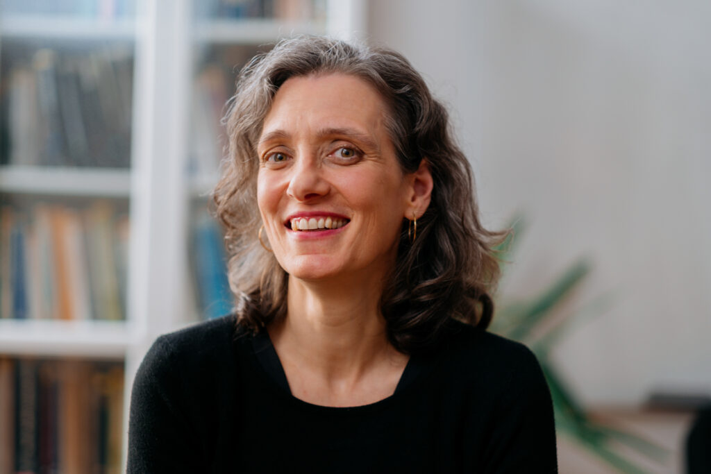 A woman with curly, dark hair, wearing a black top, smiles at the camera in a sunny room with bookcases behind her.