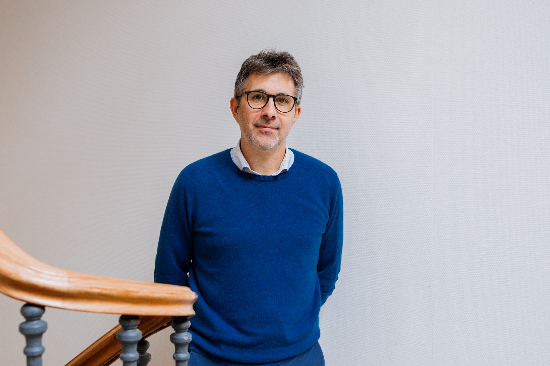 A man with glasses, wearing a blue sweater, stands in a stairwell.