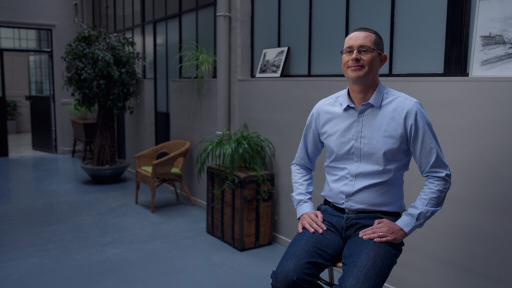 A man with glasses, wearing a pale blue shirt and gray pants, sits on a stool in a retro-industrial-style office.