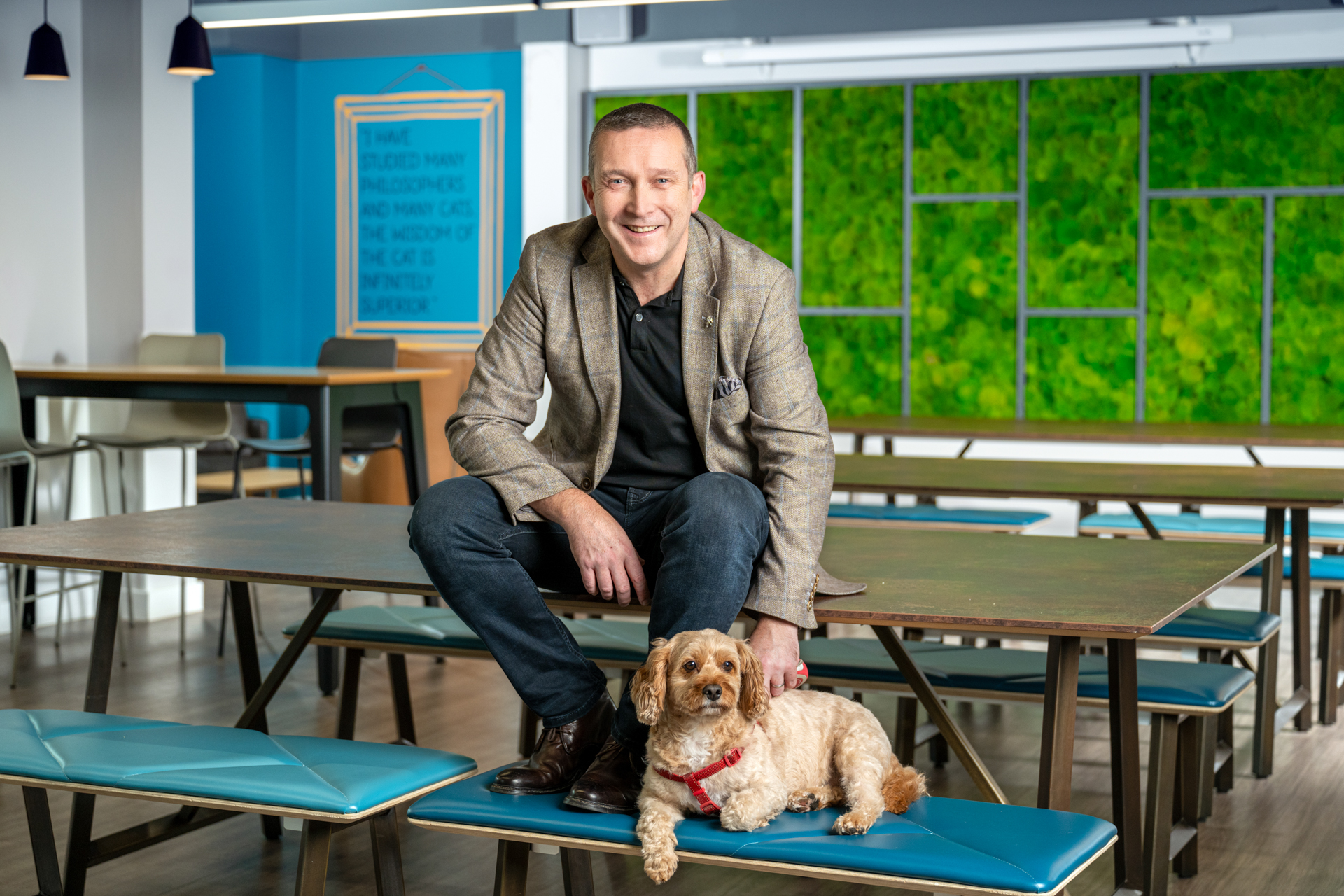 Smiling man sitting on top of a table with a light brown dog with curly hair sitting on a bench at his feet.