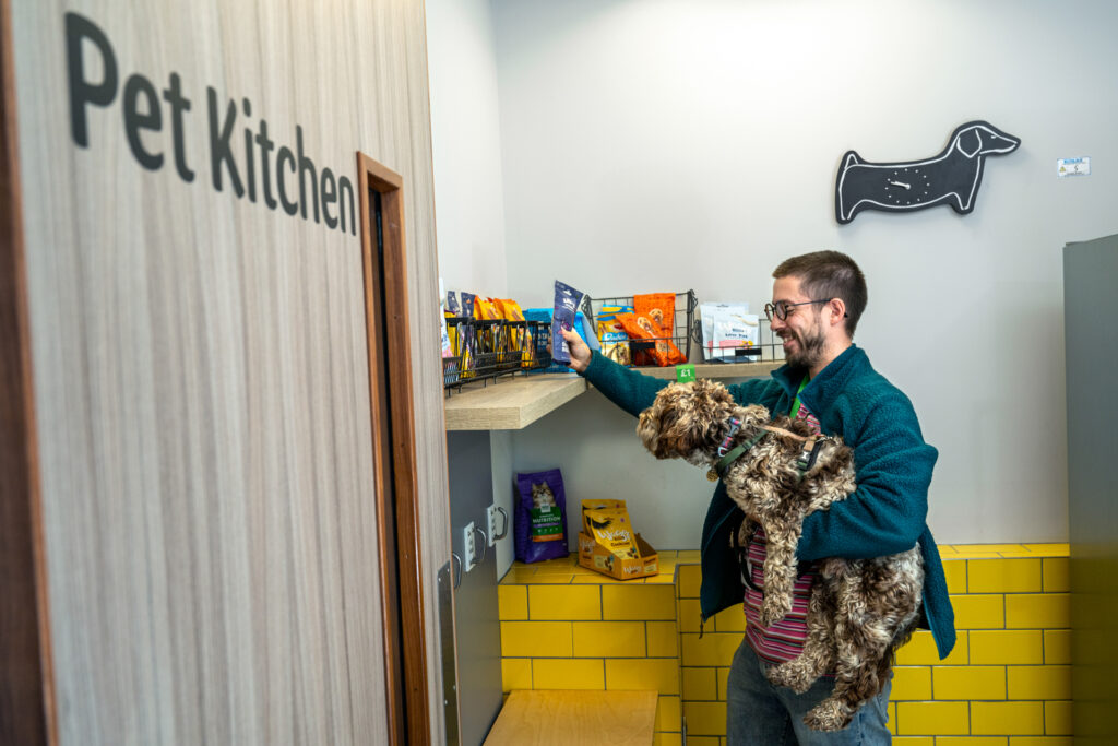 A man holding a dog in a room with a sign that says “Pet Kitchen” on the wall. The man and dog are looking at a selection of doggy treats on a shelf.