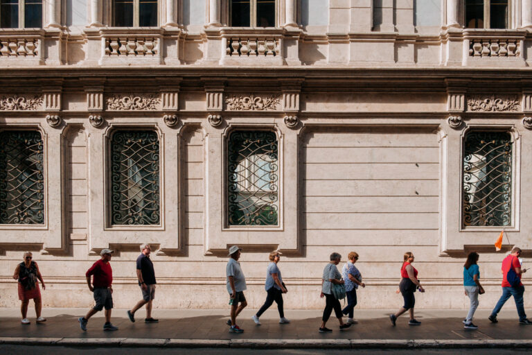 A line of tourists, following a leader with a flag, on a city sidewalk with an ornate building behind them.