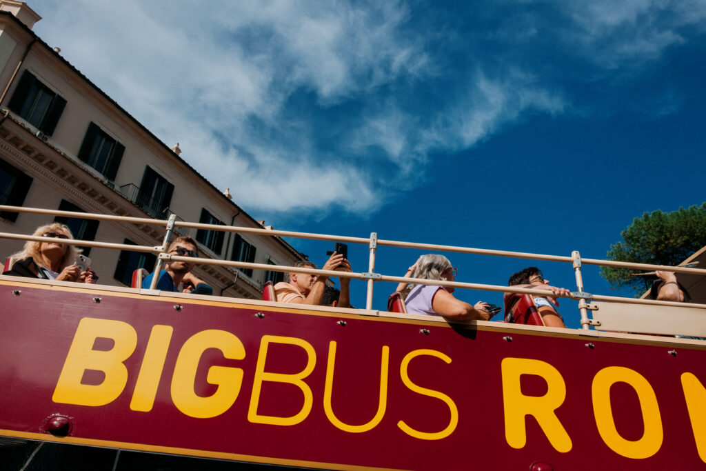 Travelers on an open topped tour bus, as seen from below, are looking at the sites and taking photos with their smartphones.