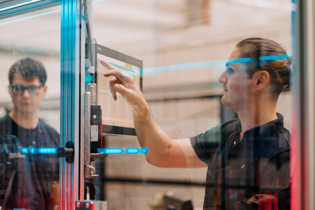 Seen through reflective glass, one man on the right touches a computer screen while another looks at the movements in the machine.  