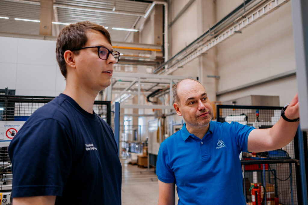 A young man in a blue T-shirt and wearing glasses, on the left, with an older man in a light blue polo shirt in a factory setting.