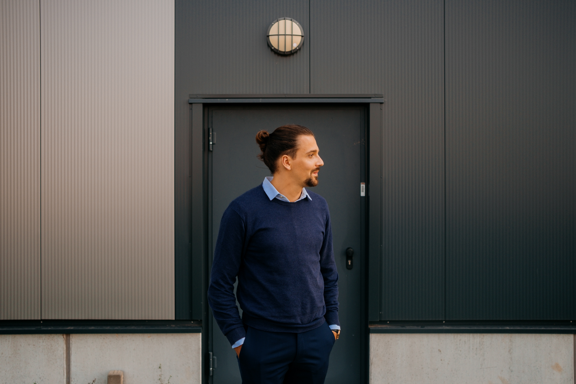 Young man with hair pulled back in a blue sweater in front of a corner of a building.