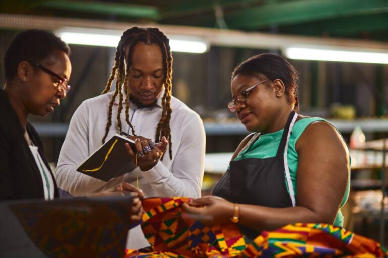 A business owner is talking with two other people while holding a piece of fabric and a man makes notes in a book.