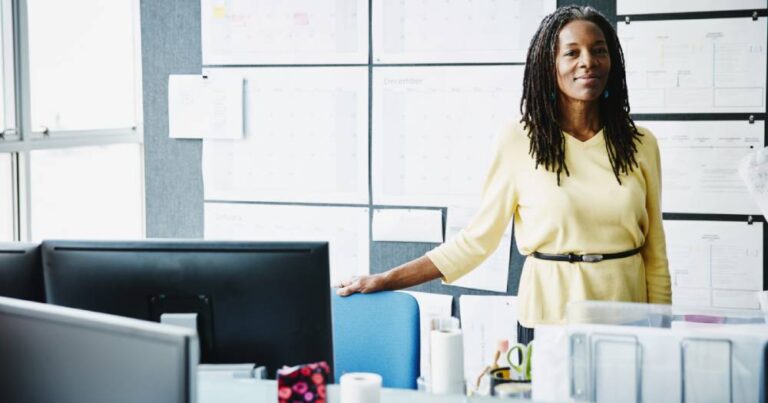 Confident businesswoman standing behind an office desk, in front of wall with paper notes in the background