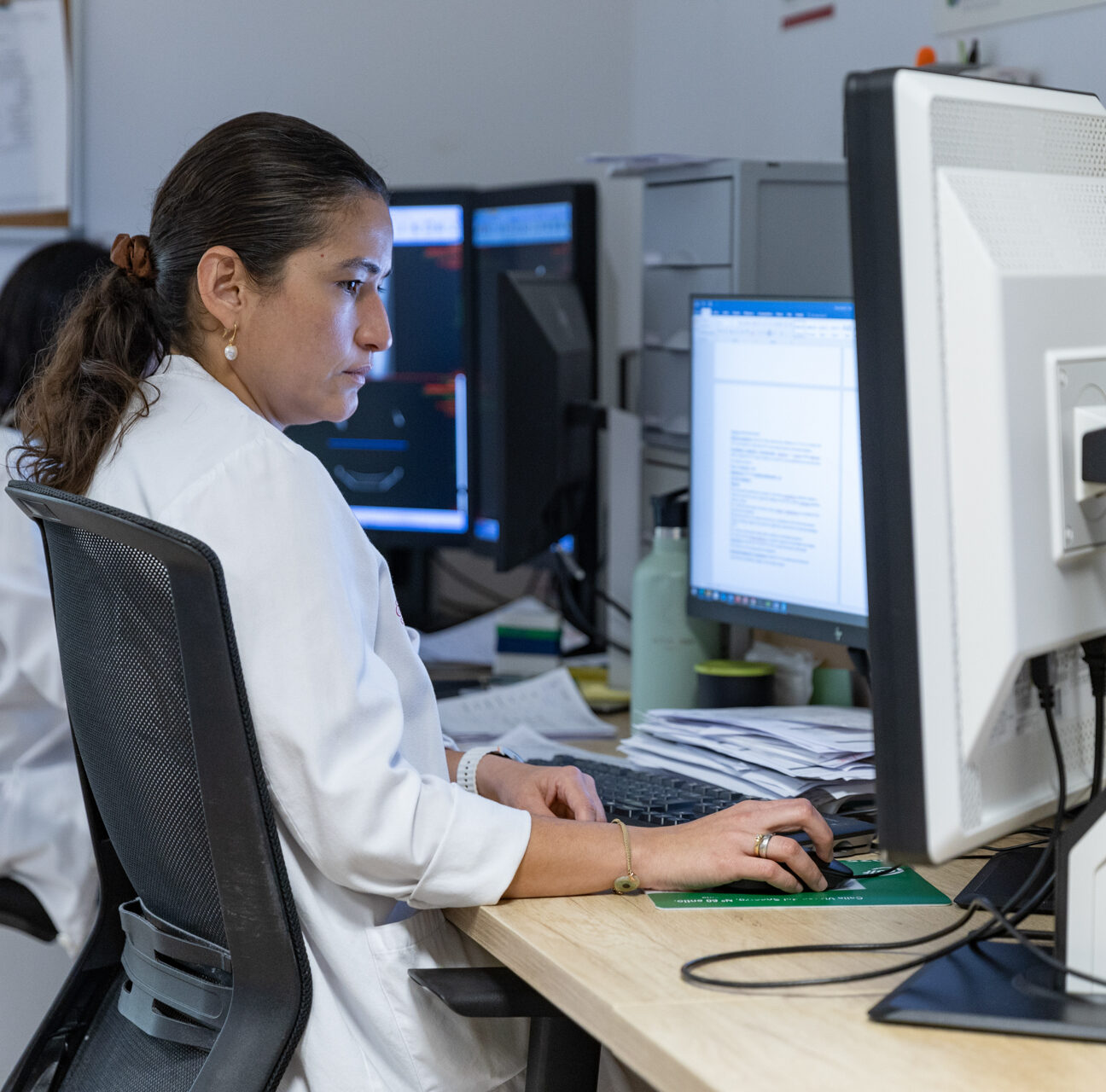 A woman in a white lab coat works at a computer.