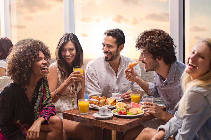 Cinco invitados a bordo de un barco de Costa se sientan en una mesa pequeña y disfrutan de bebidas de frutas y platos de comida.
