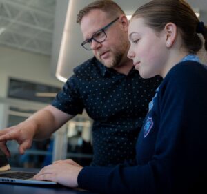Homem adulto branco aponta para computador enquanto menina branca olha