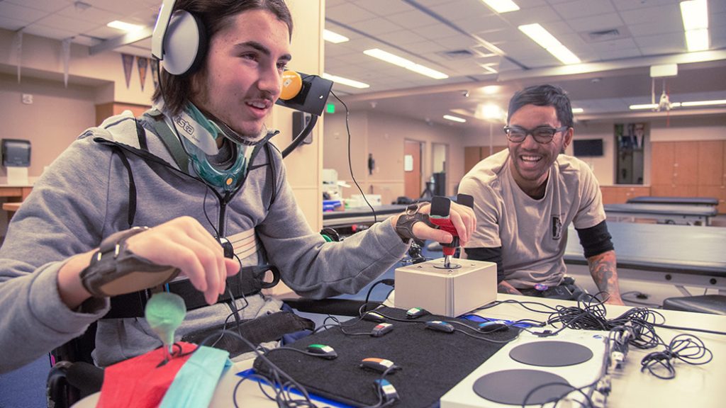 Dos jóvenes juegan a través del control Xbox Adaptive con diferentes botones e interruptores externos. Hay un hombre a la izquierda con auriculares y controlando el dispositivo con un joystick que sostiene en su mano derecha. Hay un hombre a la derecha sonriendo y sosteniendo un pequeño controlador inalámbrico que tiene un solo botón. Están sentados en una mesa con monitores, teclados y cables a su alrededor.