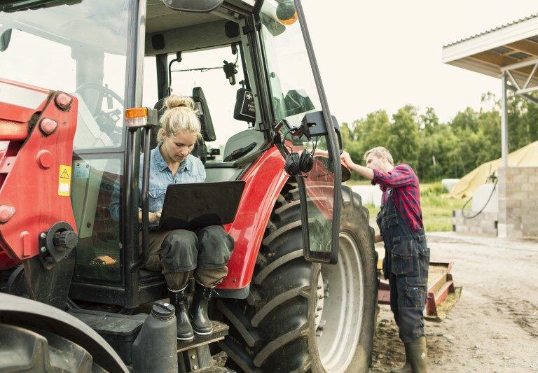 Una mujer trabaja en una laptop sentada en un tractor mientras un hombre revisa el vehículo