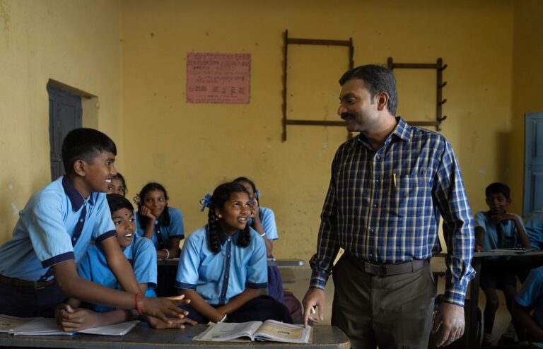 Un profesor con una camisa azul a cuadros interactuando con estudiantes con uniformes azules en un salón de clases