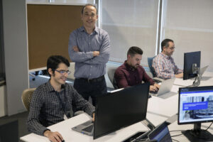 A man stands with arms folded behind three workers at computer desks