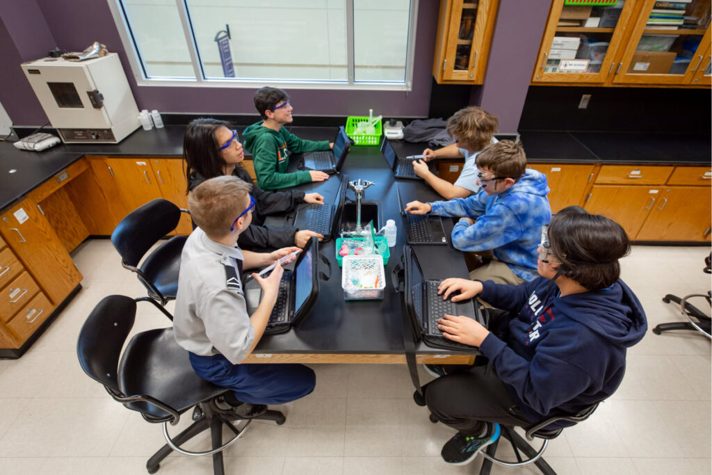 Imagen de un grupo de estudiantes sentados alrededor de una mesa en un aula comparando notas.