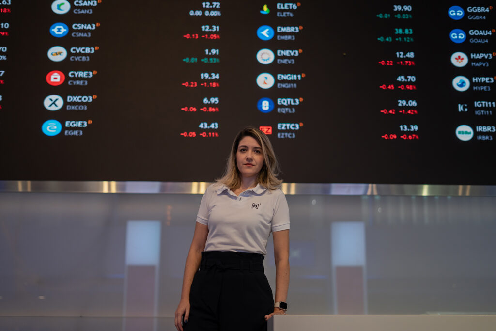 Christianne Bariquelli stands in front of a huge black screen that displays ticker symbols in vivid colors.