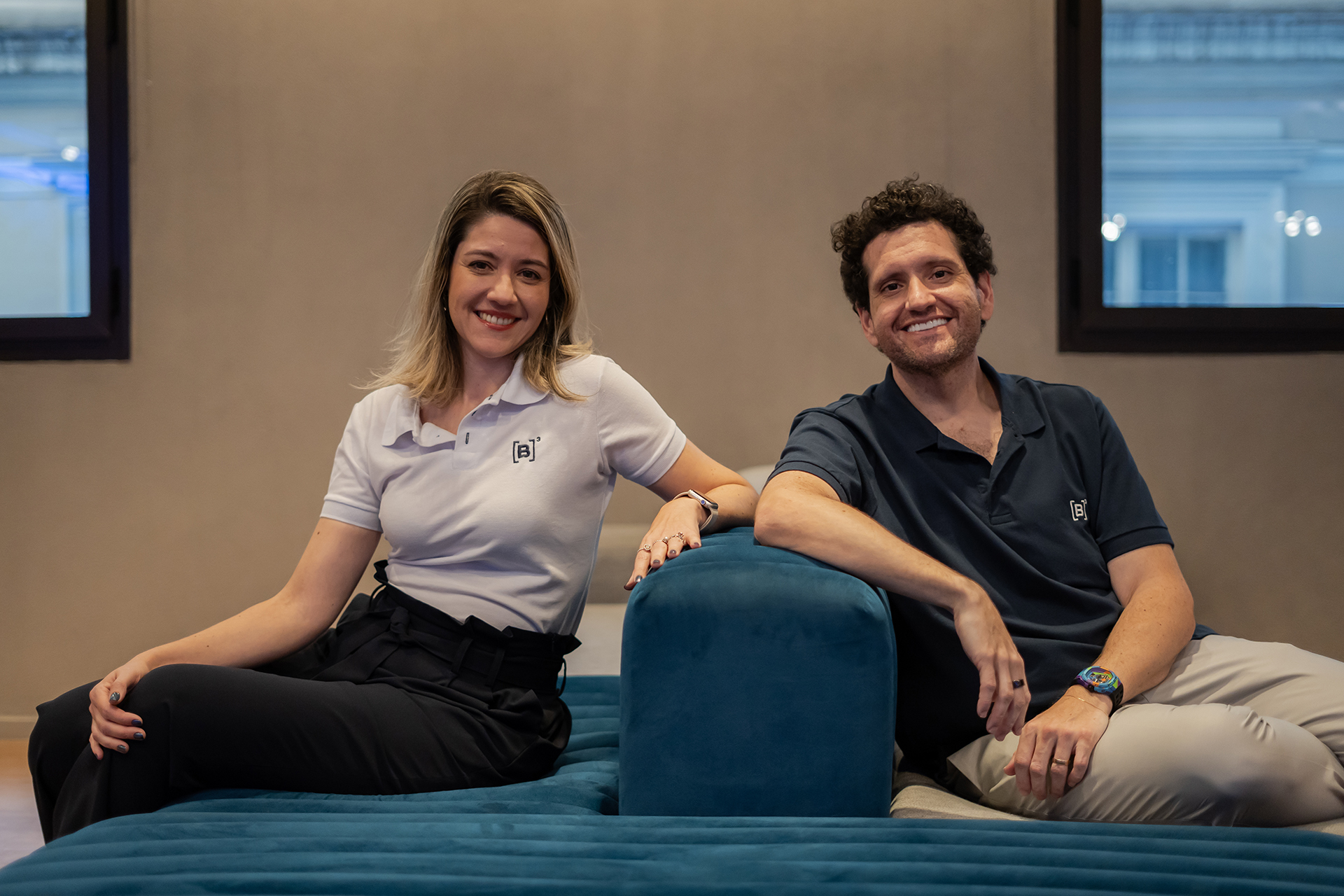 Christianne Bariquelli e Marcos Albino Rodrigues sentados em um sofá, de frente para a câmera. Ambos estão sorrindo e vestindo camisas polo com o logotipo da B3.