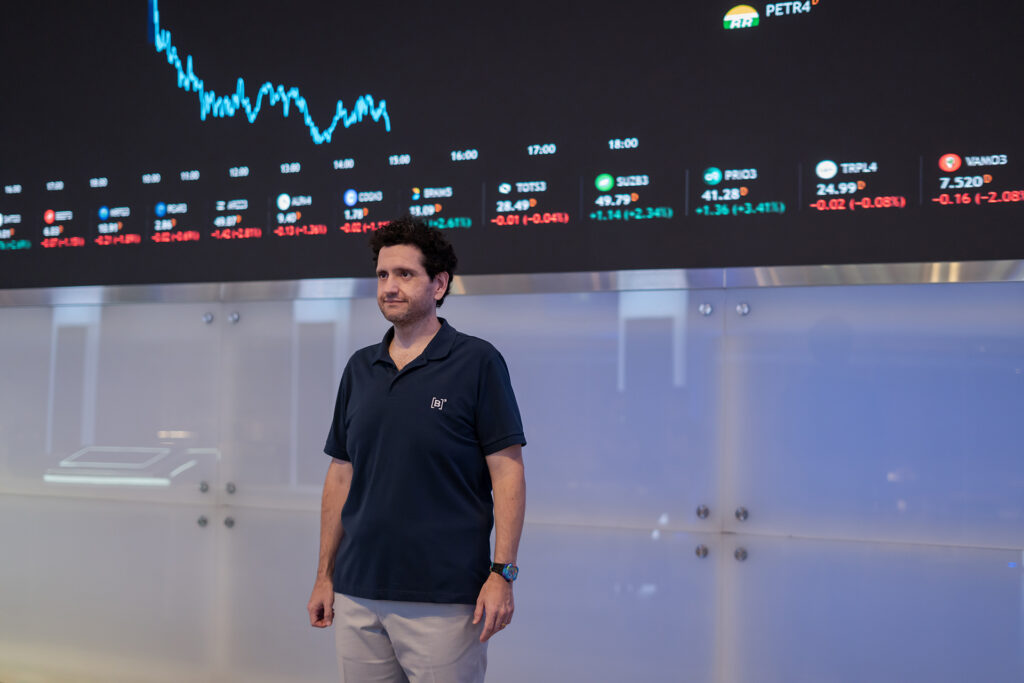 Marcos Albino Rodrigues stands in front of a huge black screen that displays ticker symbols and graphs in vivid colors.