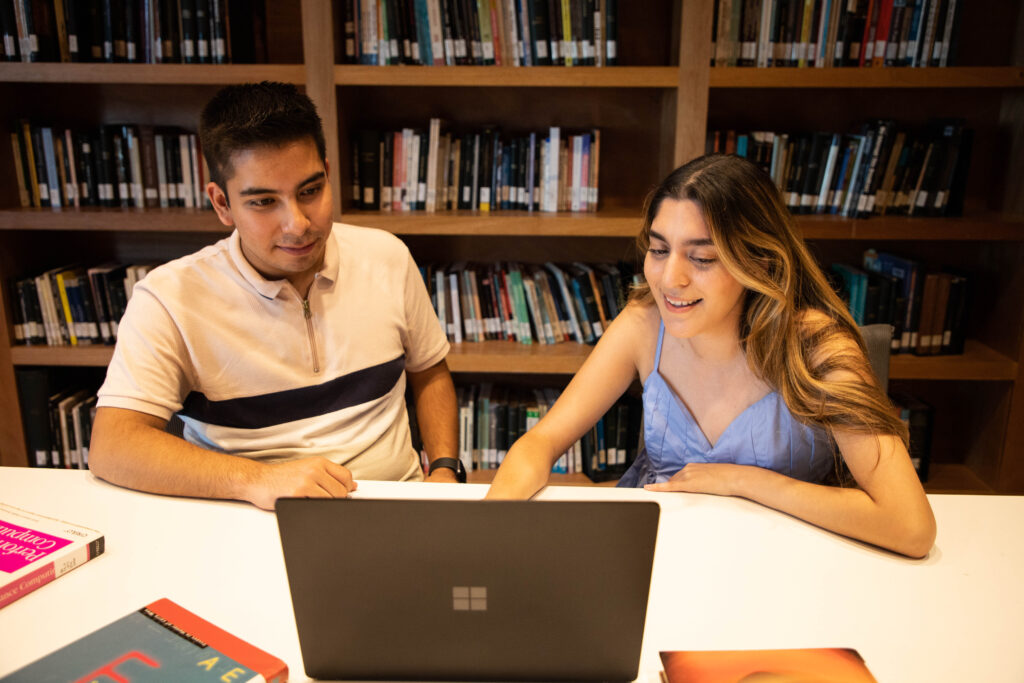 Un estudiante y una estudiante están sentados en un escritorio mirando una computadora portátil, con estantes llenos de libros detrás de ellos