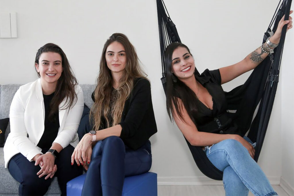 Tres mujeres sentadas juntas, una en un sofá, otra en una otomana, otra en una silla hamaca 