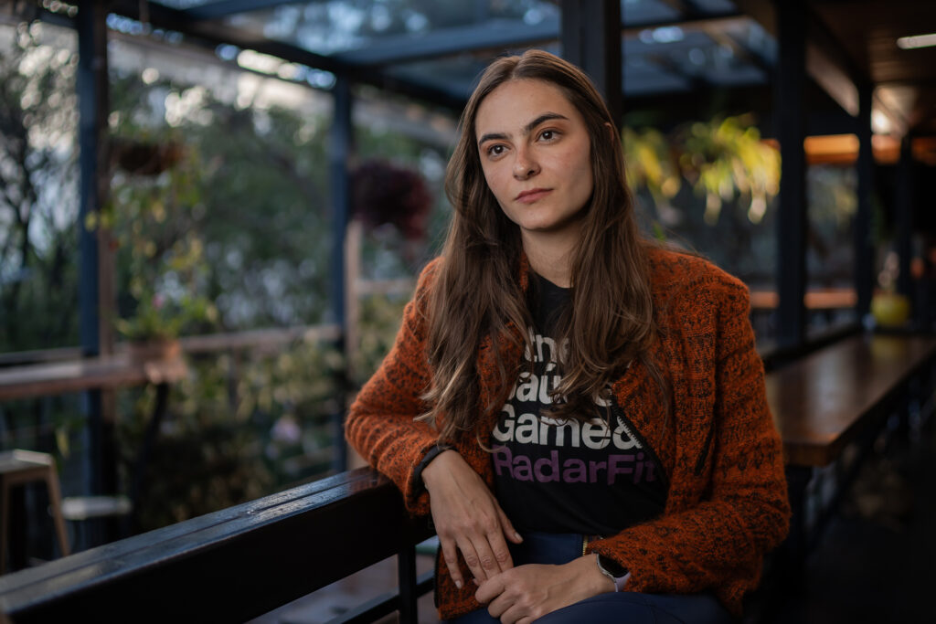 A woman leans against a rail with plants in the background