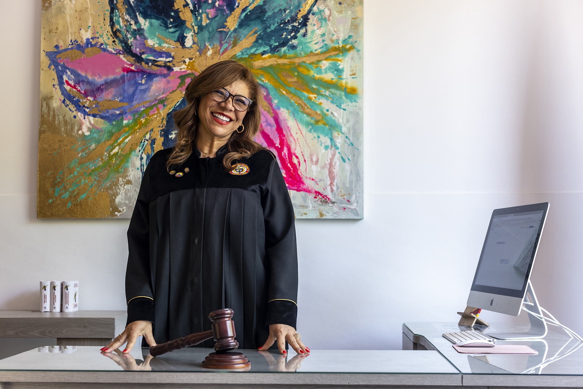 Una jueza con ropas judiciales está de pie en un escritorio con un mazo frente a ella, detrás de ella hay una pintura colorida y a su lado una computadora.