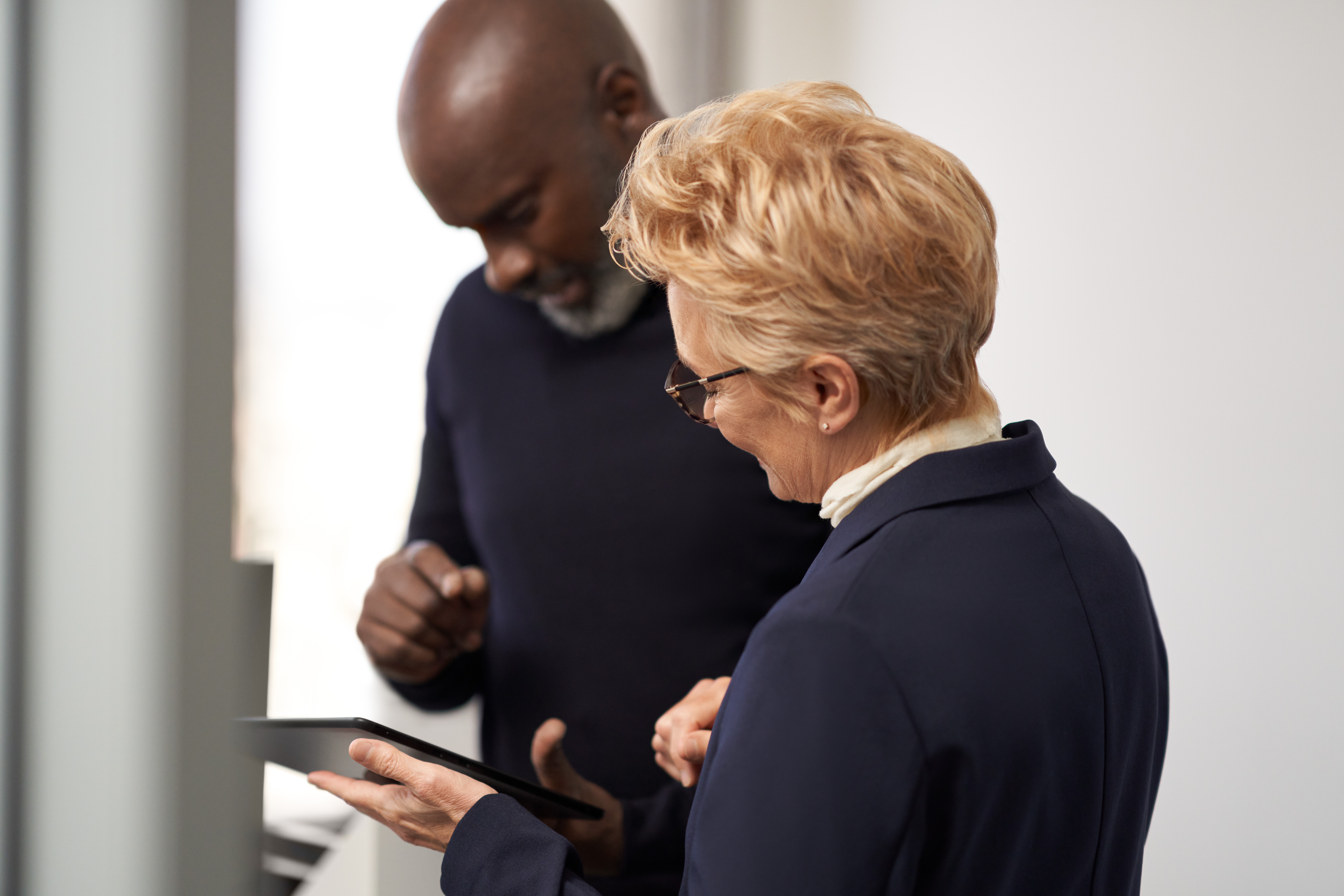 Duas pessoas em ambiente corporativo observam juntas a tela de um tablet. Uma delas segura o dispositivo enquanto a outra acompanha atentamente o conteúdo, sugerindo uma interação colaborativa e focada. A imagem transmite uma atmosfera profissional, de troca de ideias e uso de tecnologia no trabalho.