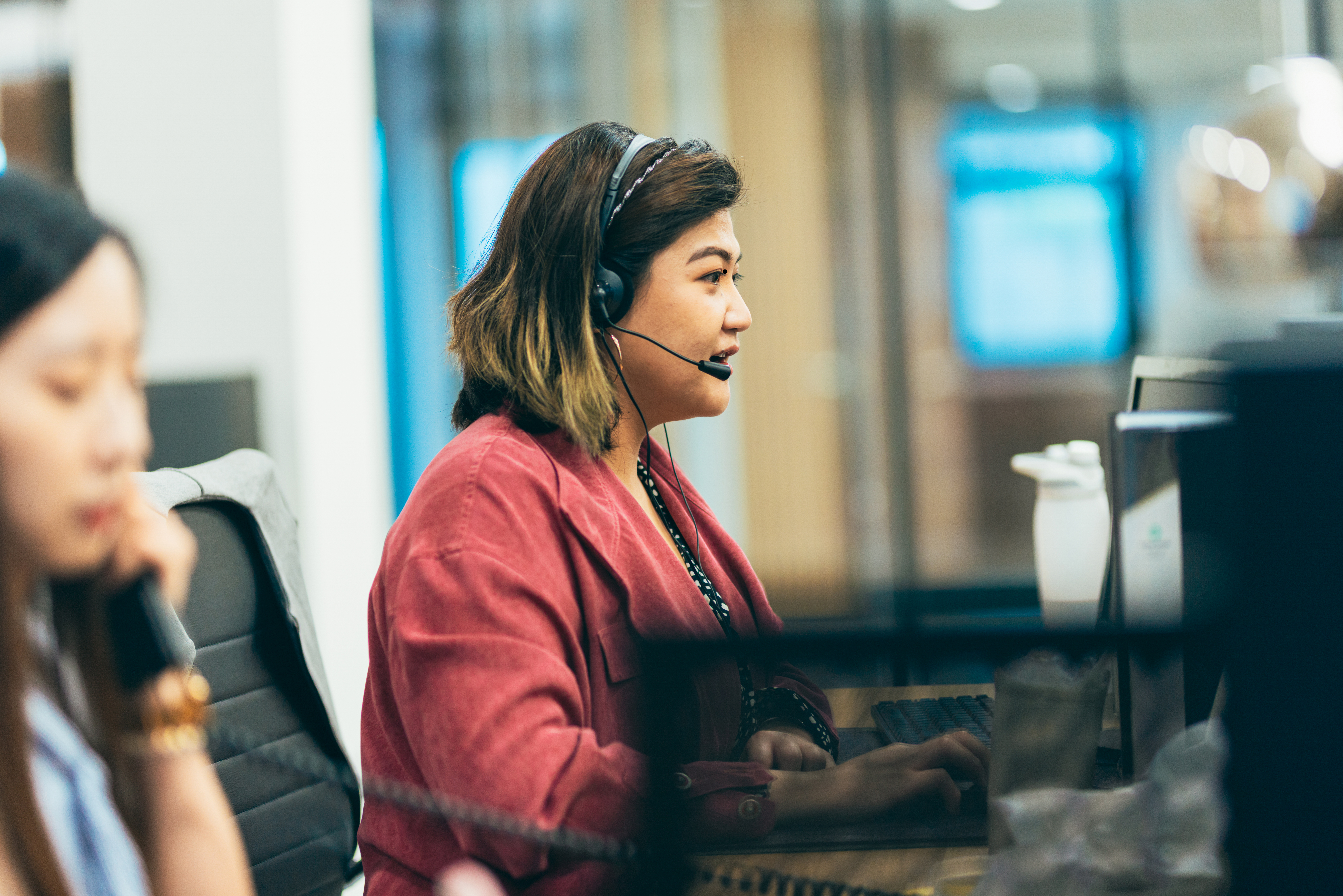 Fotografia de um ambiente corporativo, com uma profissional em destaque sentada diante de um computador e utilizando um headset com microfone. Ela veste um blazer em tom avermelhado e aparece de perfil, concentrada em um atendimento ou conversa. Ao fundo, há divisórias de vidro e telas desfocadas. No primeiro plano, outra profissional aparece parcialmente e fora de foco, também utilizando um headset. A composição transmite um contexto de atendimento, suporte ou central de operações em um escritório moderno.