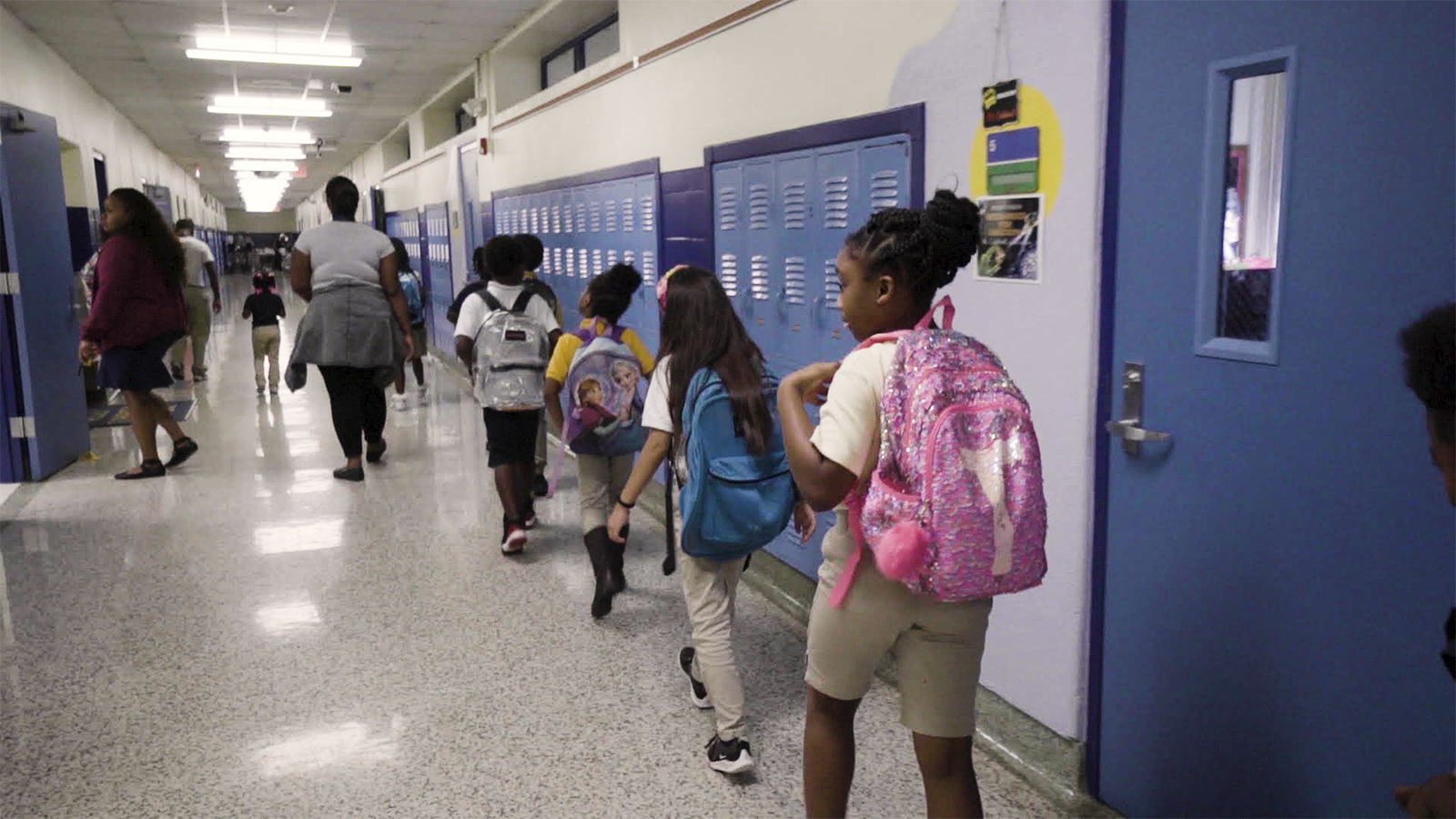 Four children, seen from the back, walk in a line inside a school hallway, next to a set of lockers.