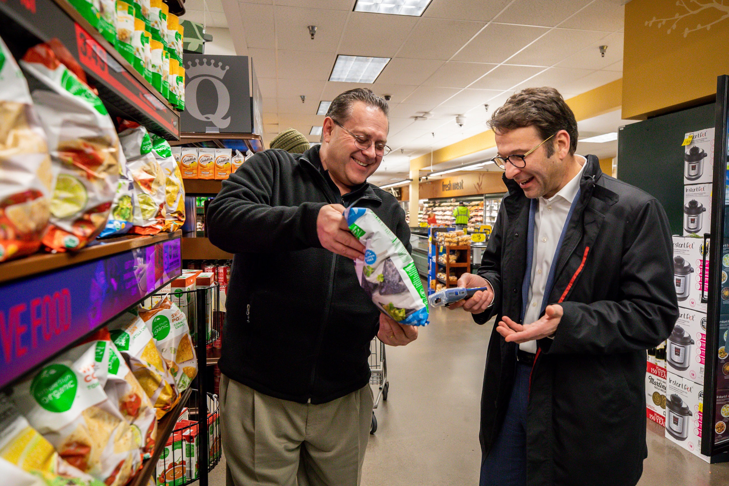 One man holds a bag of chips inside a store while a second man aims a pricing scanner at the bag.