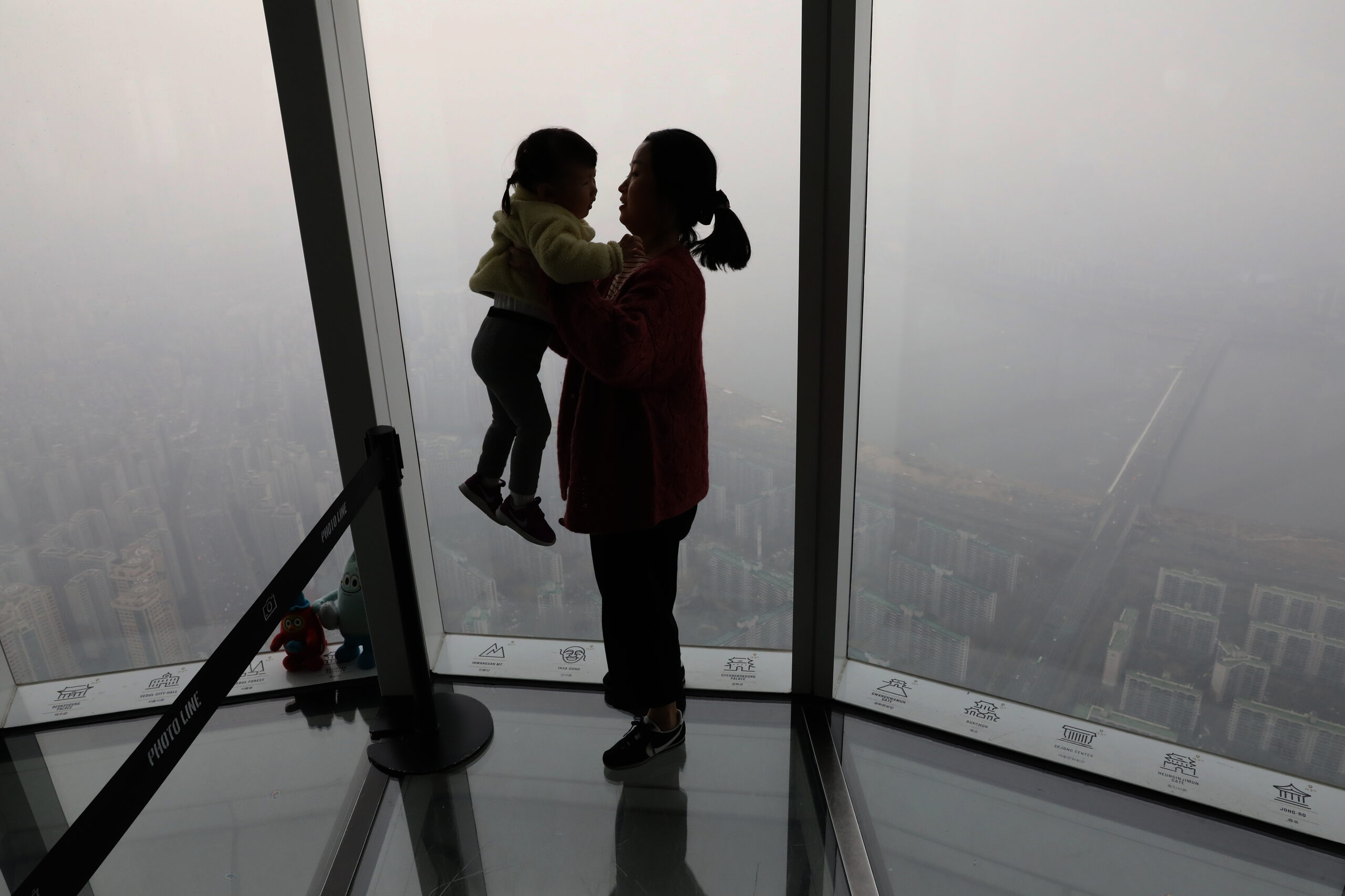 The silhouette of woman holds a small child in her arms. Both are standing in front of a window on a smoggy day.