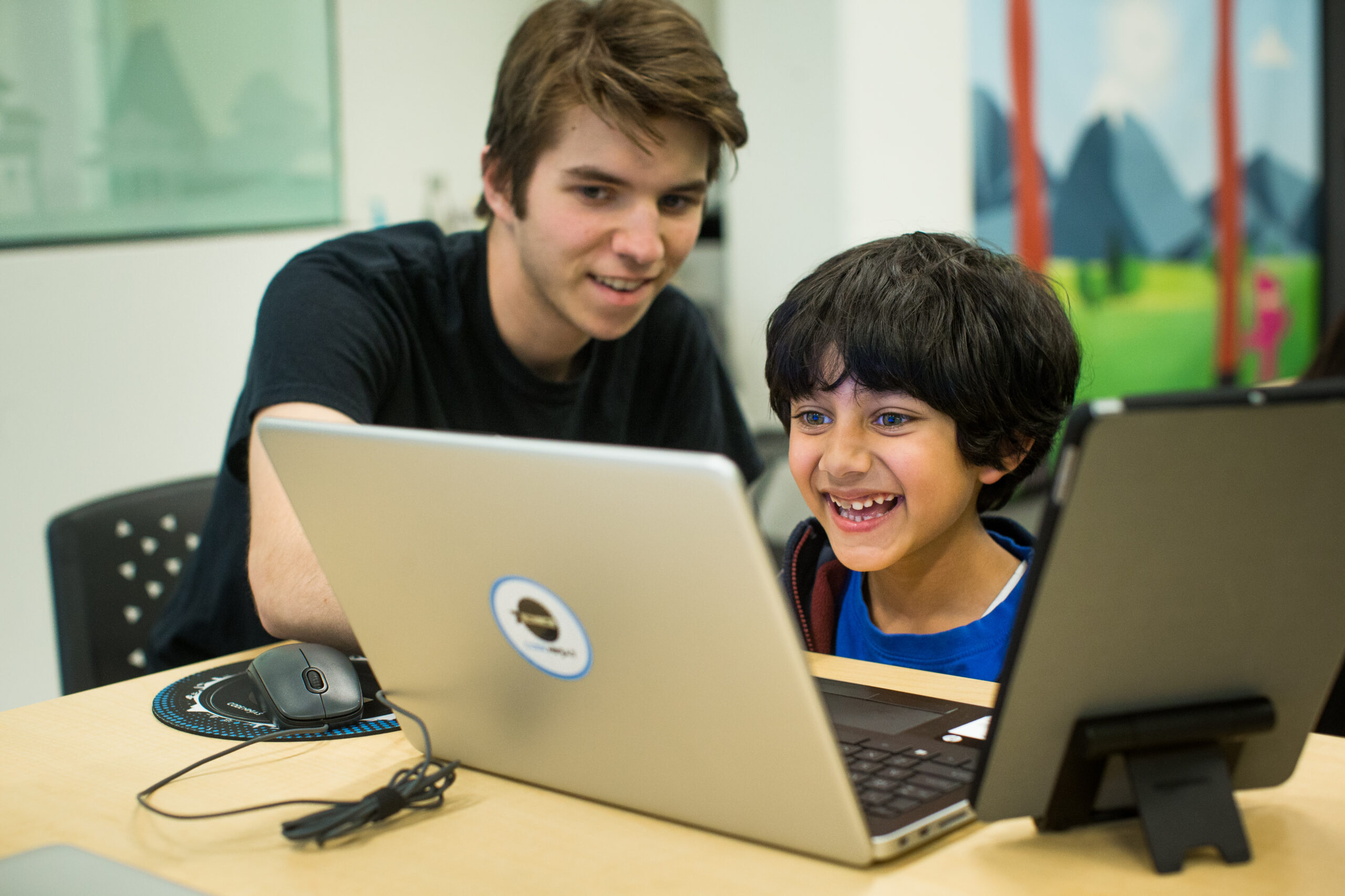 A young boy smiles into a laptop as a teenage mentor looks over his shoulder.