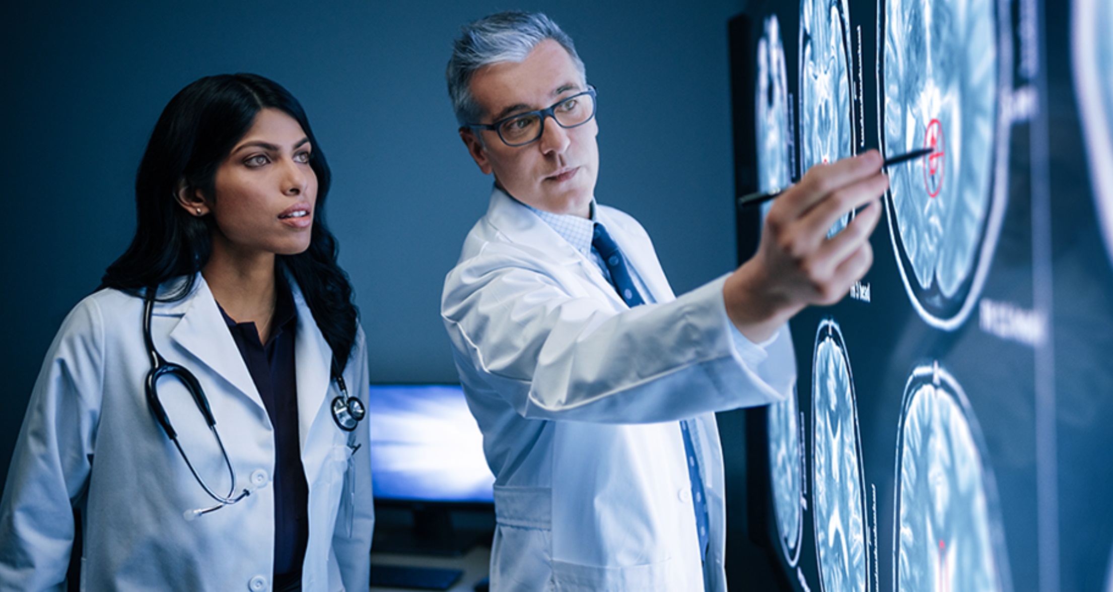 Two doctors are looking (one of them is pointing) at a large screen showing brain images.
