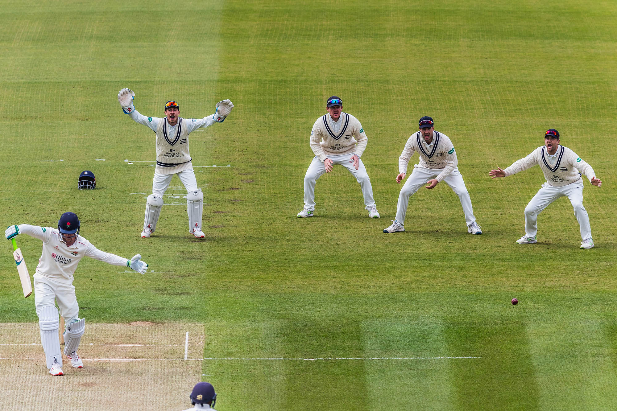 A cricket player swings and misses while four cricket players behind him react.