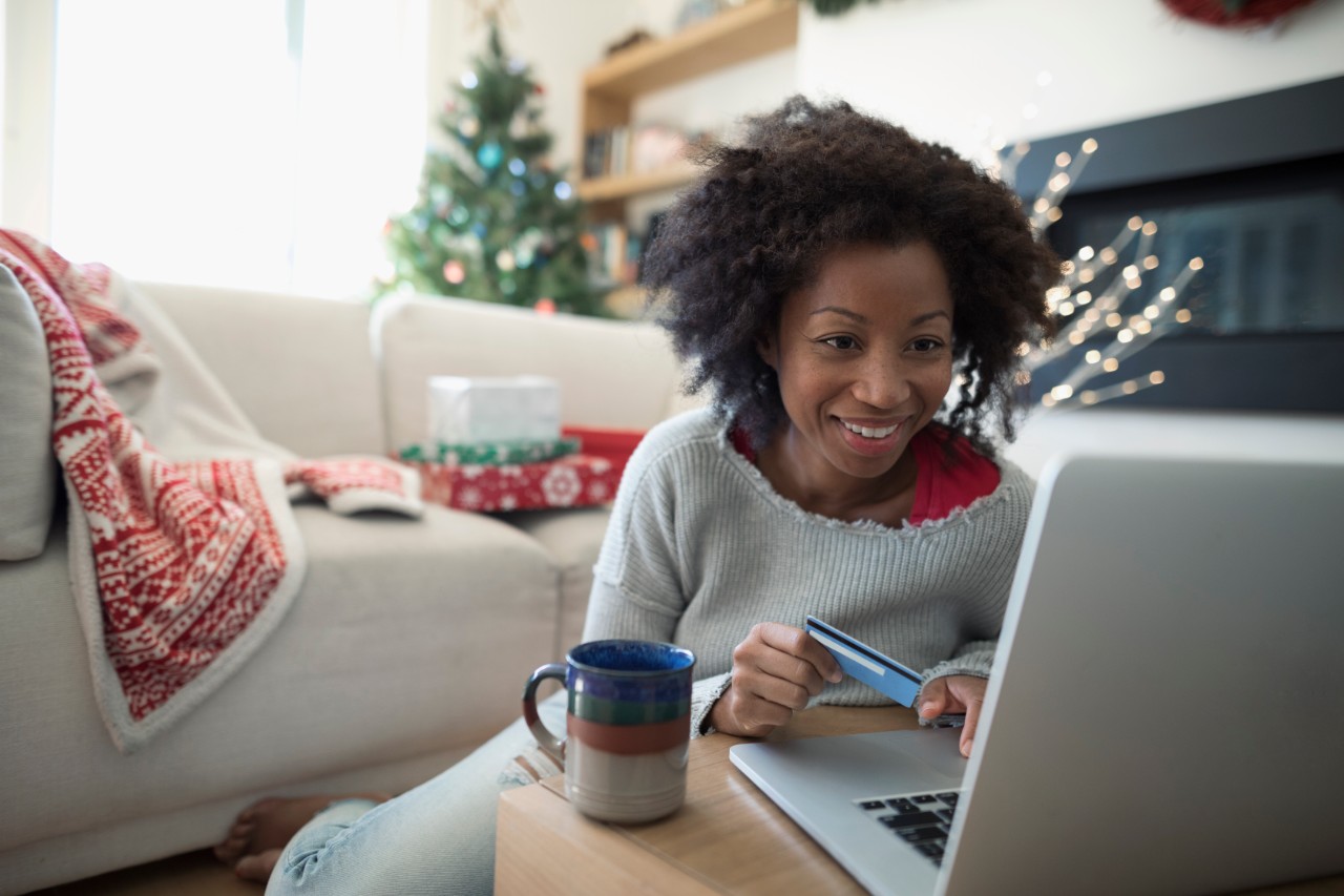 Photo of a woman sitting on the floor in a living room looking at a computer, with presents and a Christmas tree in the background.