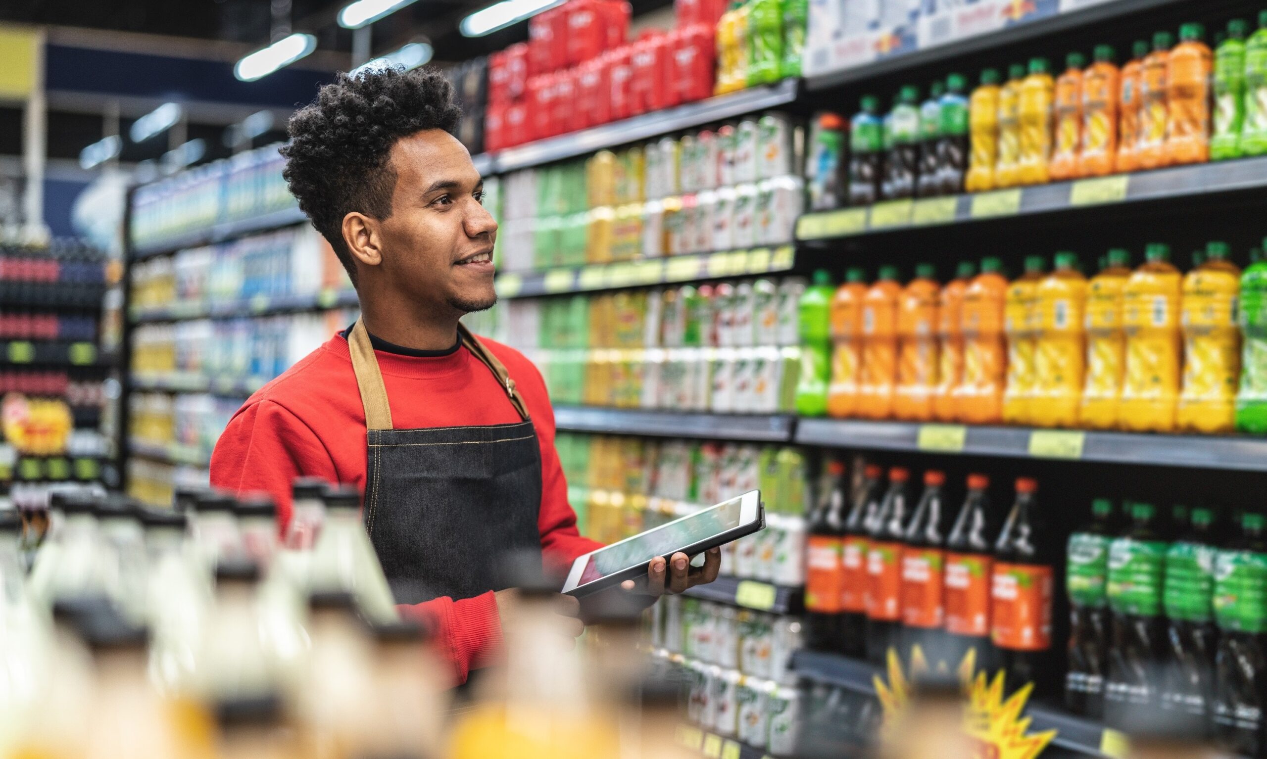 Photo of a young male grocery store employee holding a tablet and standing in front of shelves of beverages.