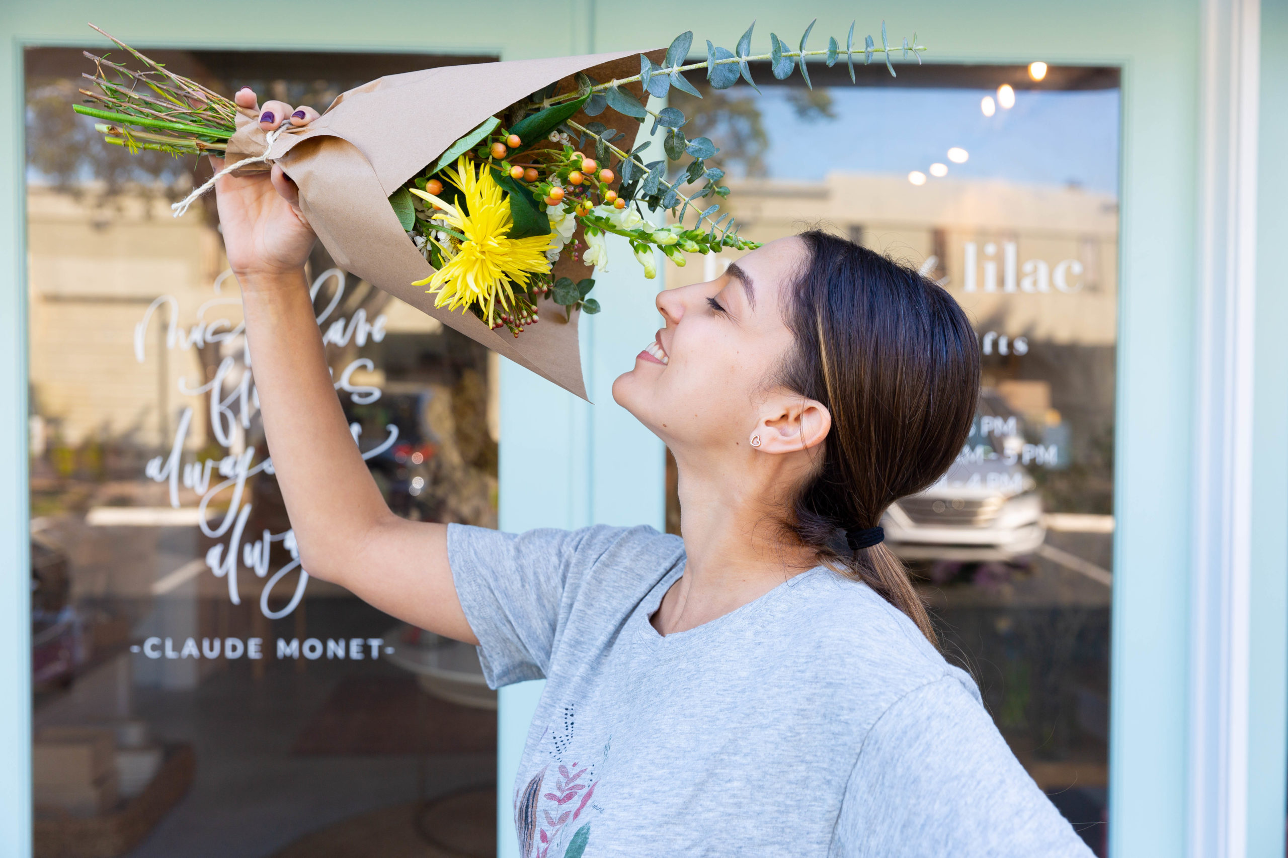 Smiling woman stands outside Miami flower shop House of Lilac, holding up a wrapped bouquet of flowers.